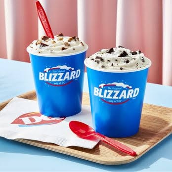 Two Dairy Queen Blizzard cups with whipped cream and toppings on a tray with DQ napkin and red spoons