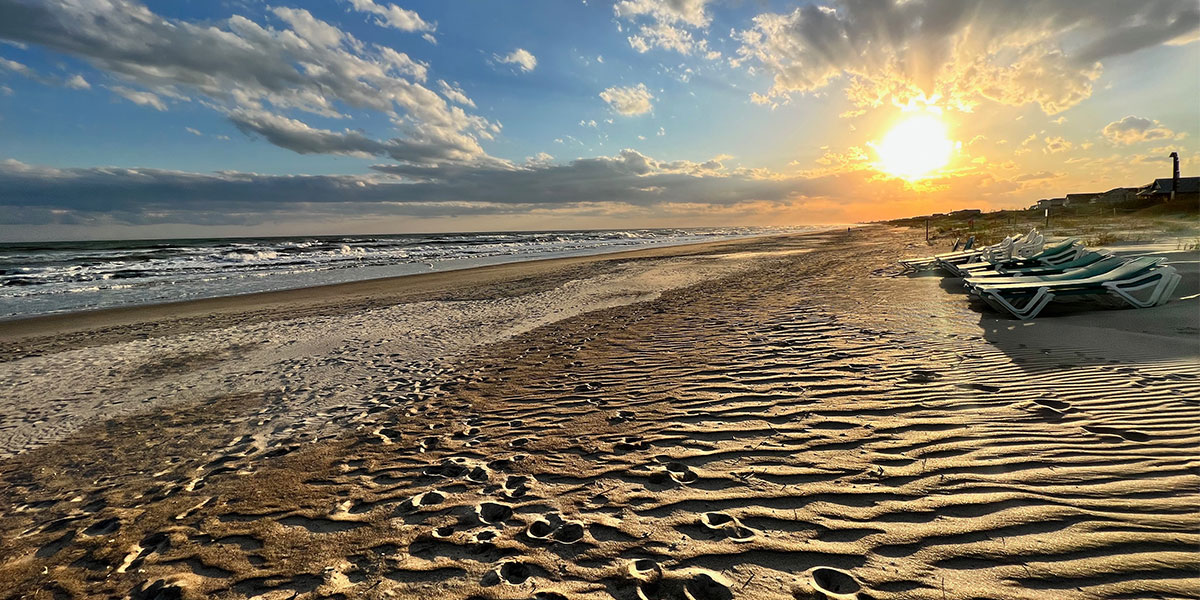 Sunrise over Atlantic Beach NC with golden light on rippled sand beach chairs and lighthouse silhouette in the distance